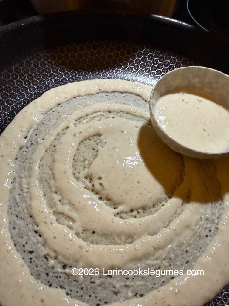 A ladle spreading batter in a circular motion to make canjeero on a frying pan.