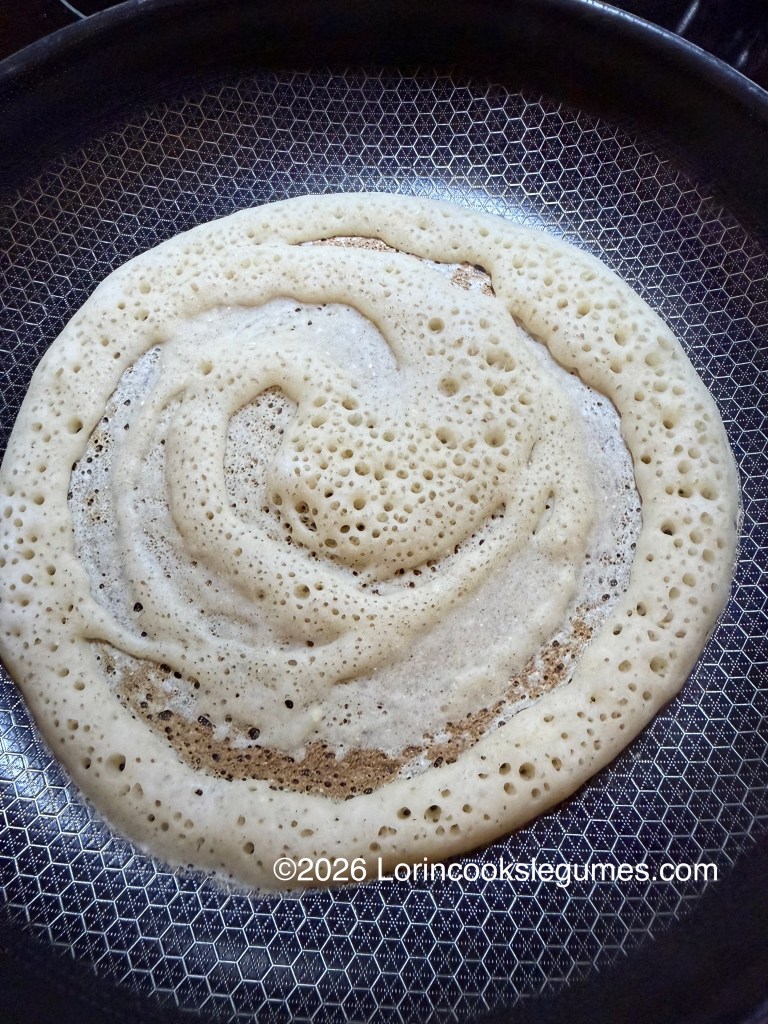 A close-up view of a crepe-like canjeero, cooking in a frying pan, showcasing bubbles forming on the surface as it cooks.
