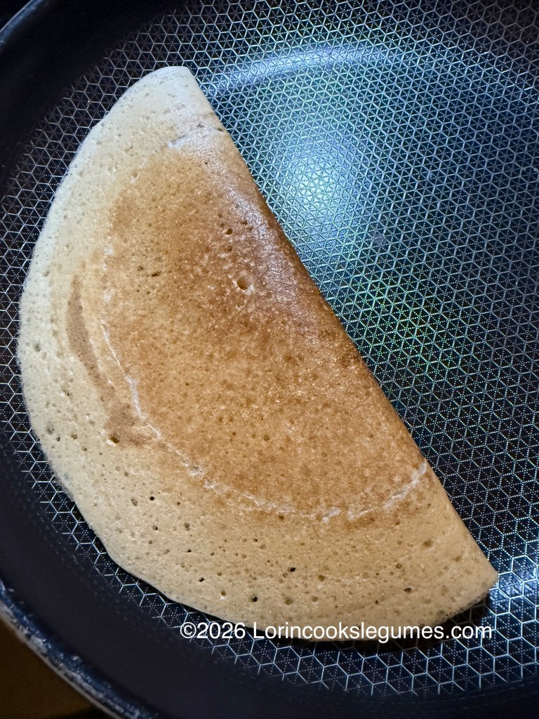 A finished canjeero, folded in half on a non-stick frying pan, showing a golden-brown surface flecked with small holes.