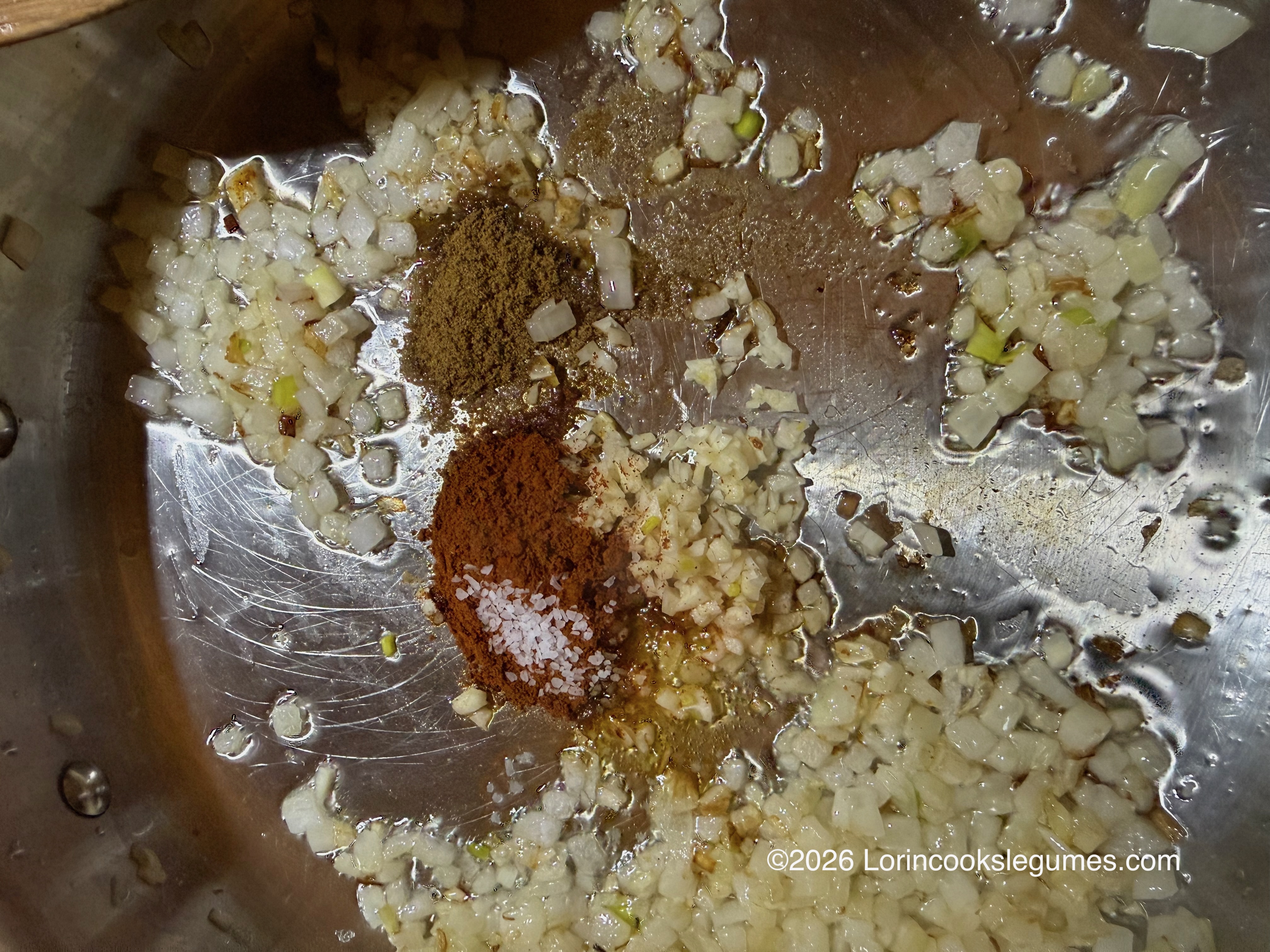 Diced onions, garlic, cumin, berber powder, and salt in a stainless steel pan. ©2026 Lorincookslegumes.com.