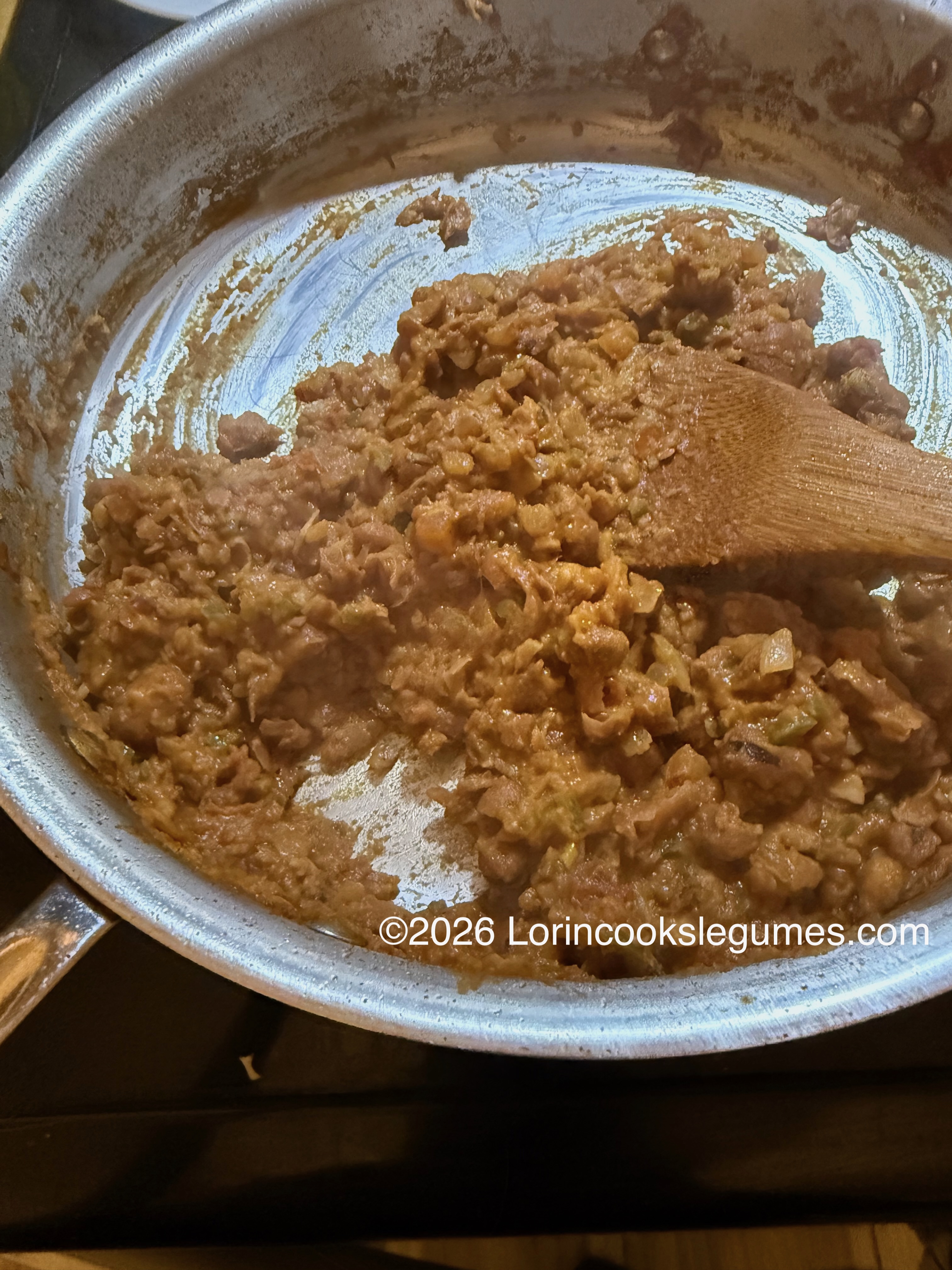 Mashed beans being stirred with a wooden spoon in a metal skillet. ©2026 Lorincookslegumes.com