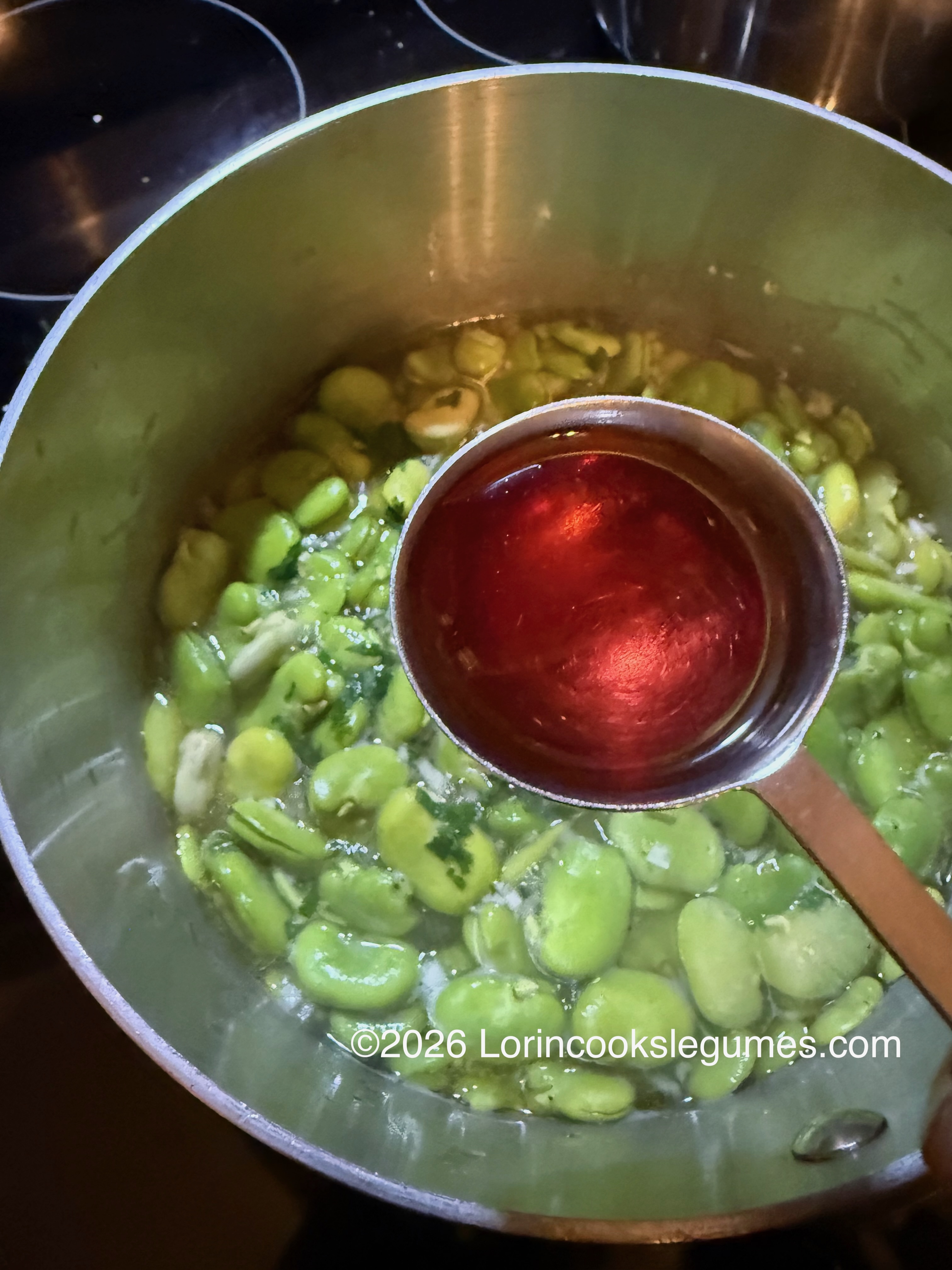 A metal spoon holding red wine vinegar above a pot of green fava beans boiling in water