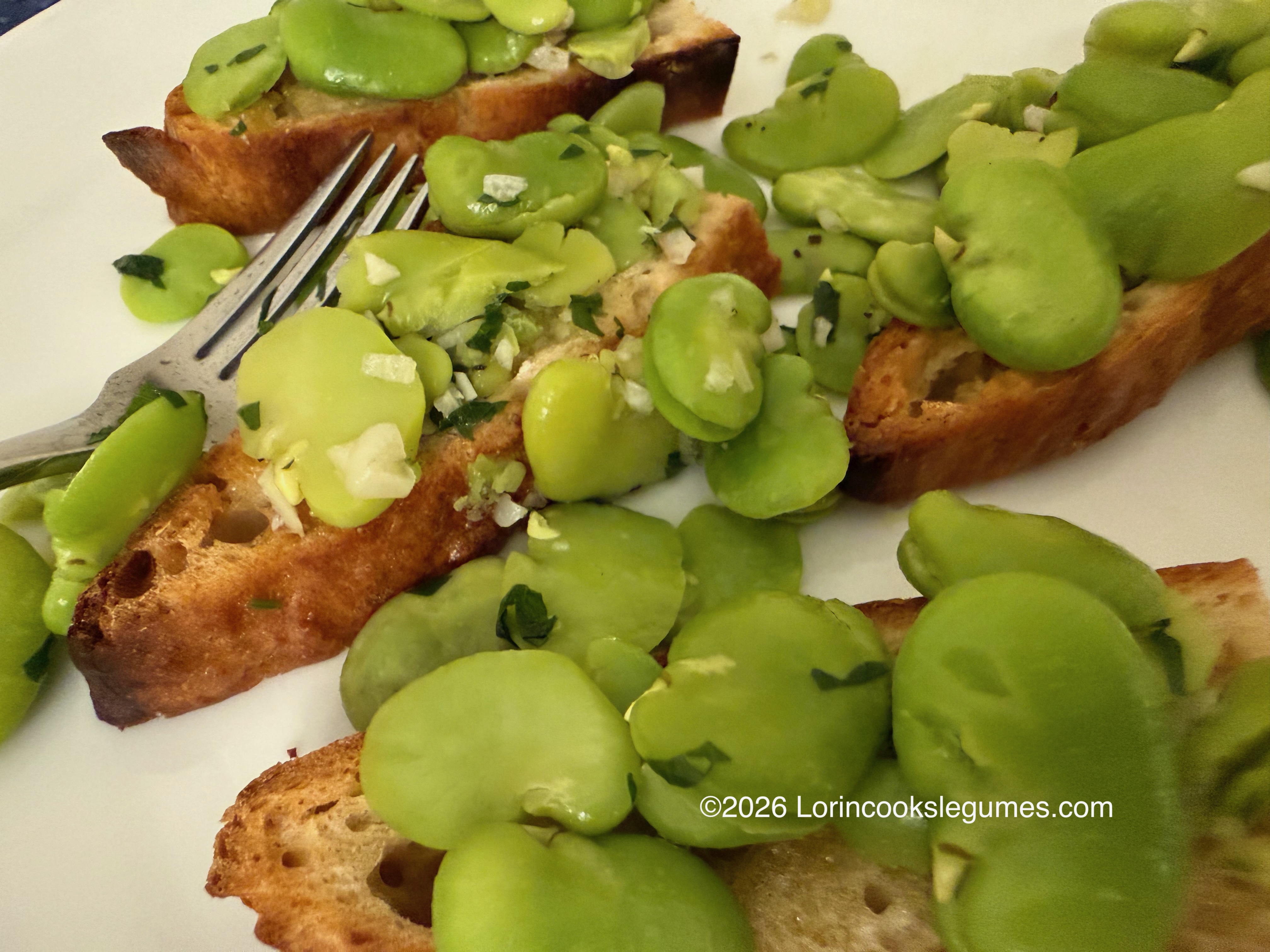 Slices of toasted bread topped with cooked broad beans, garlic, and herbs on a white plate with a fork