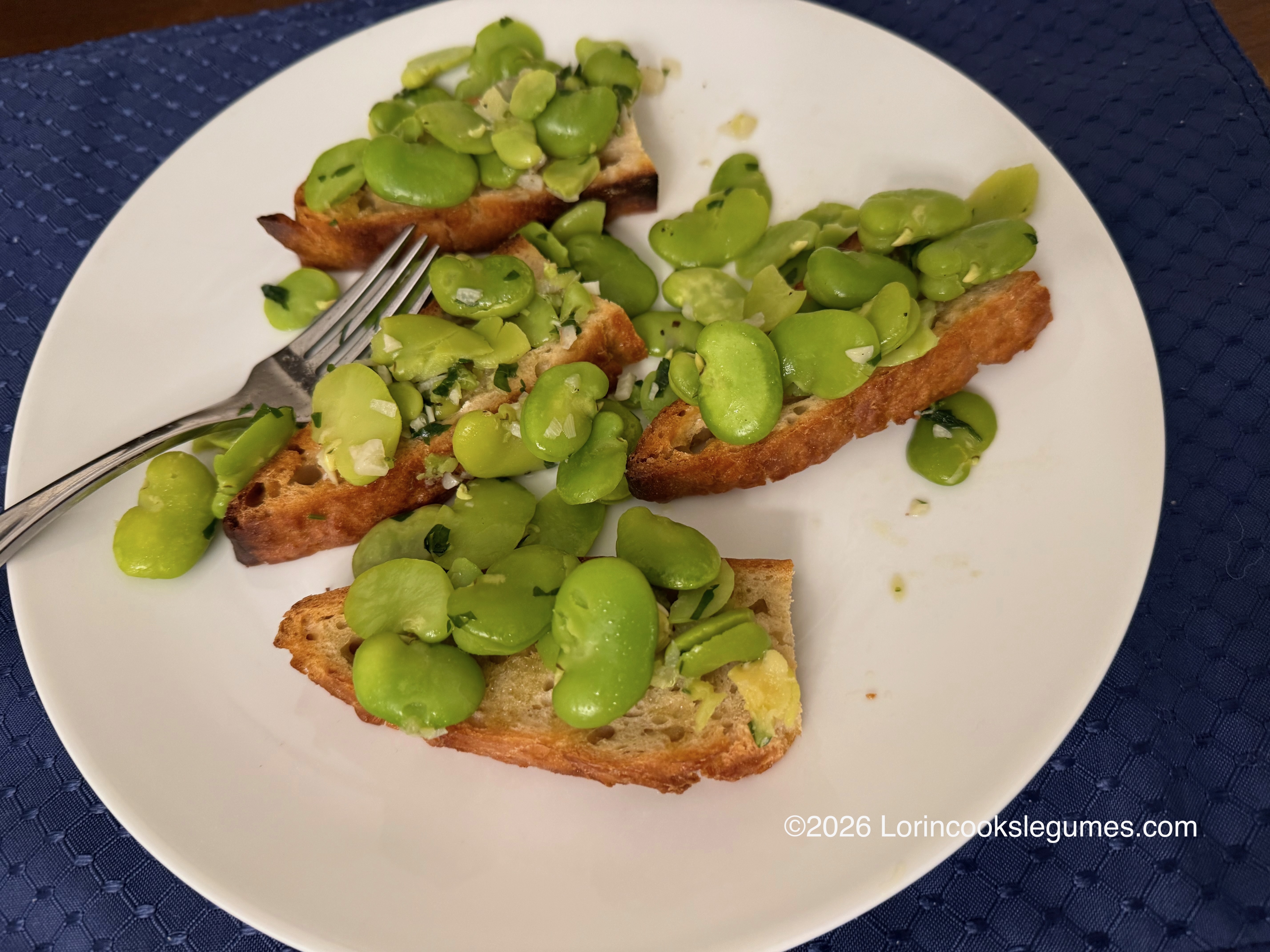 Slices of toasted bread topped with cooked broad beans and herbs on a white plate with a fork