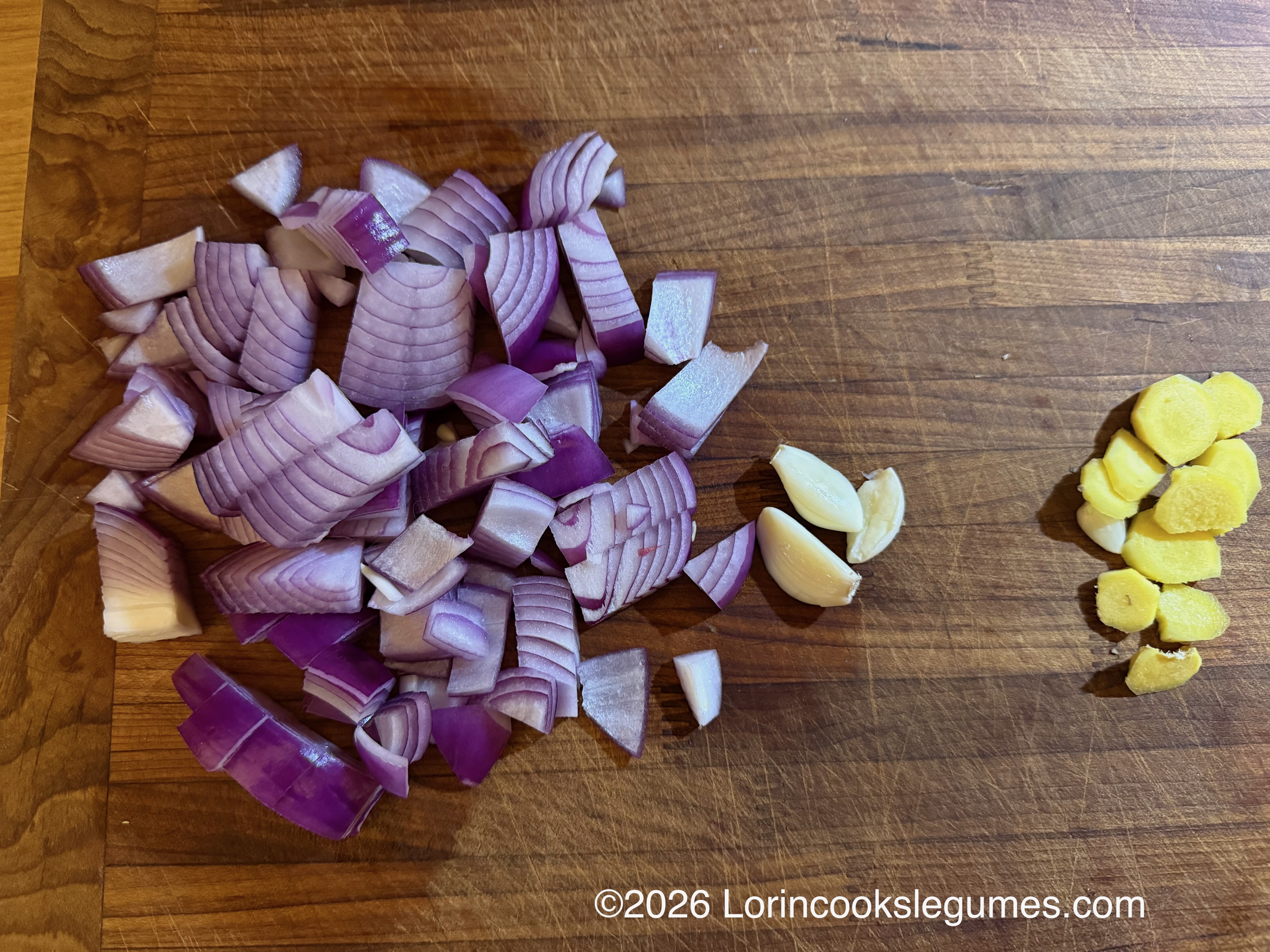 Chopped purple onion, garlic cloves, and sliced ginger on a wooden cutting board