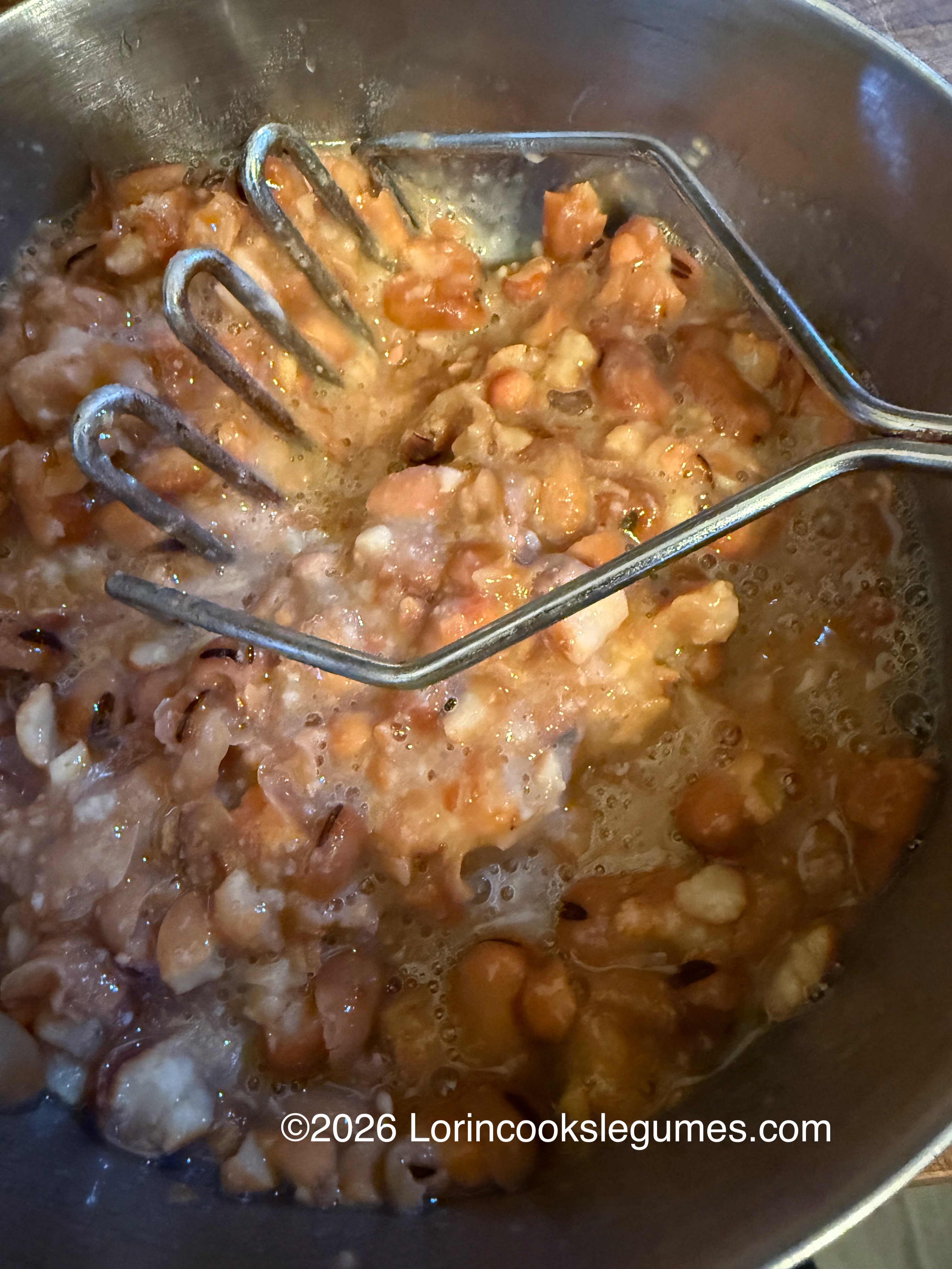 Metal potato masher mashing cooked fava beans in a metal bowl.