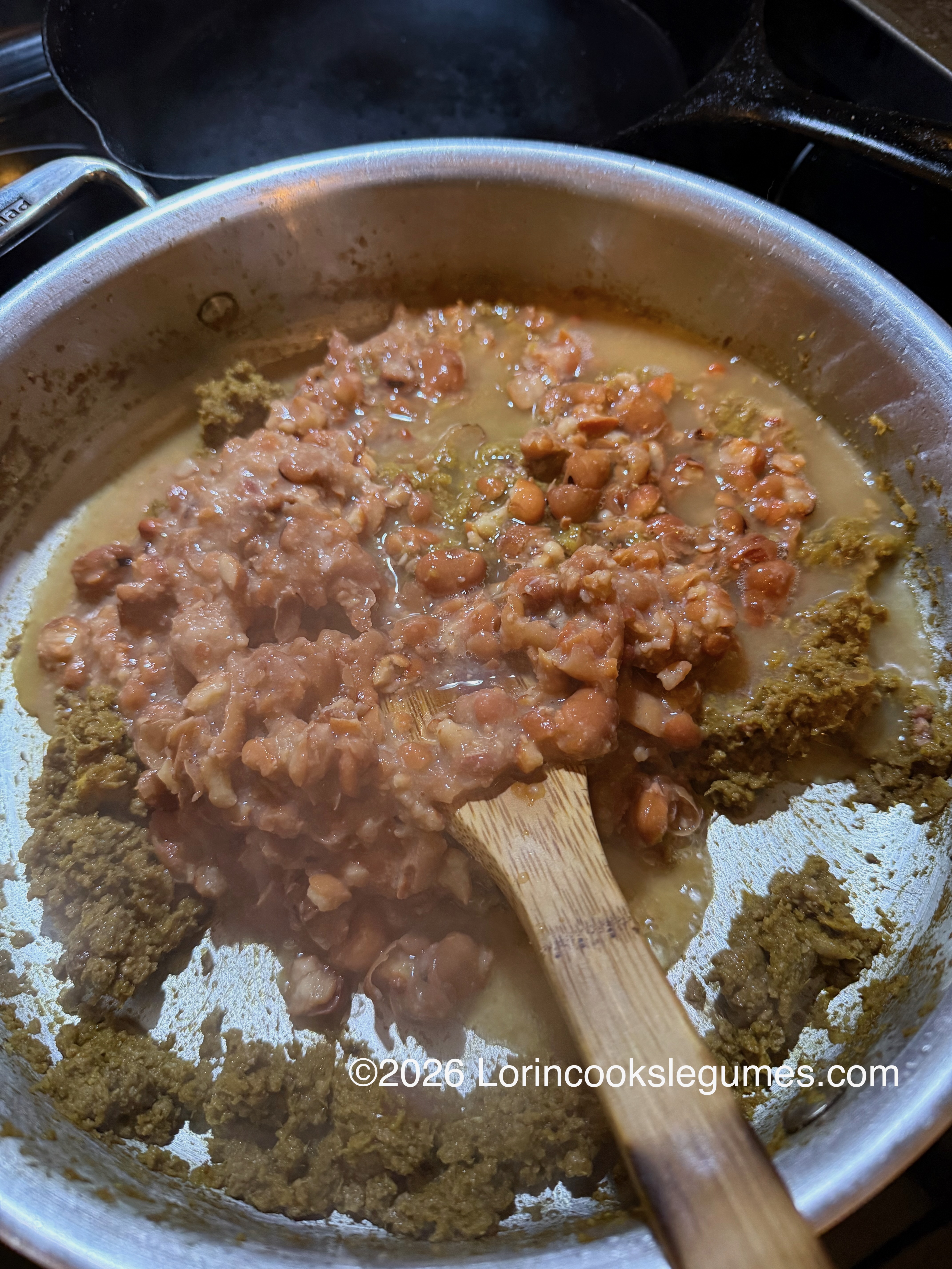 Cooked fava beans being stirred in a pot with a wooden spoon, surrounded by seasoning paste