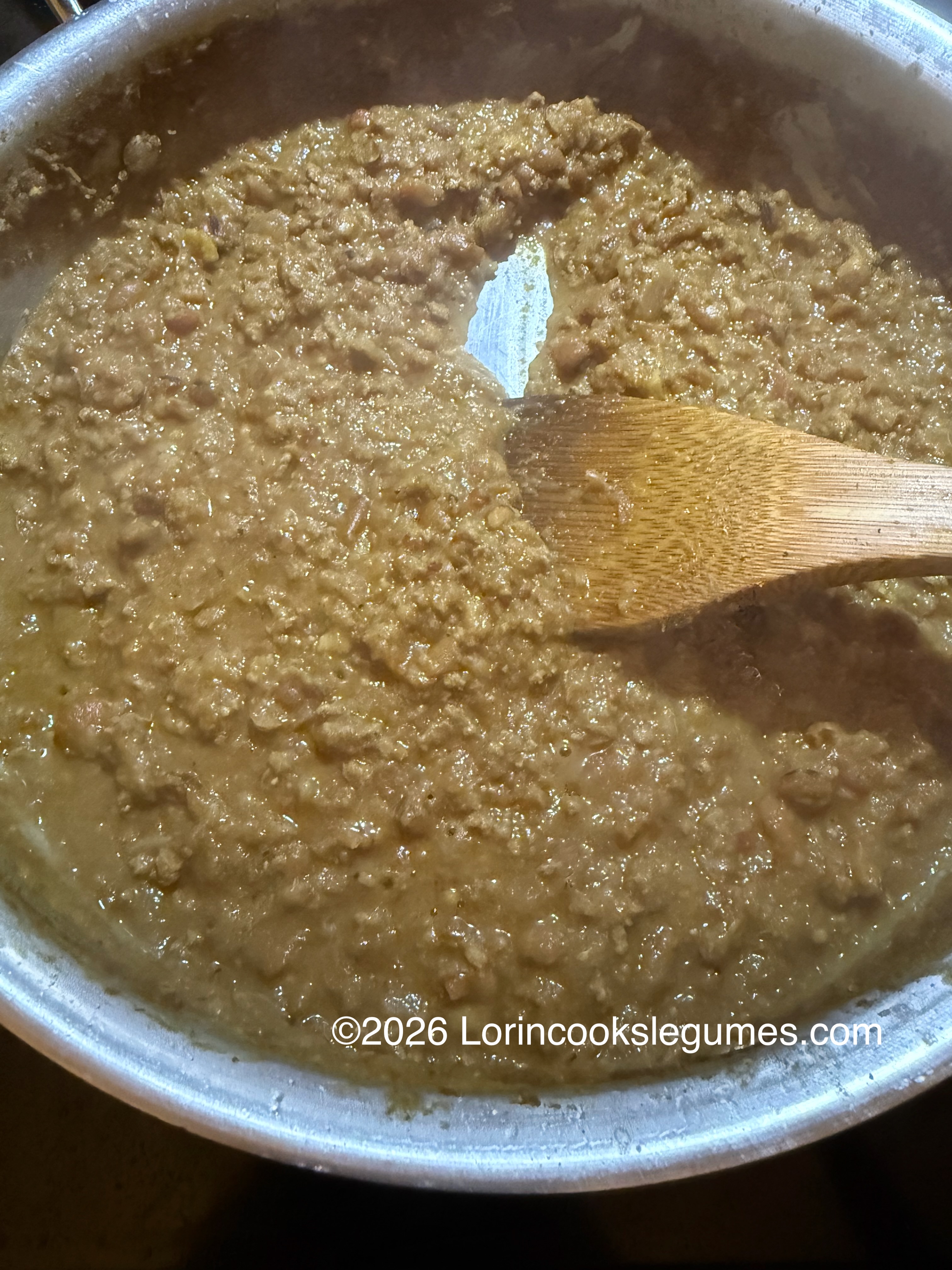 Thick brown legume stew being stirred with a wooden spatula in a pot