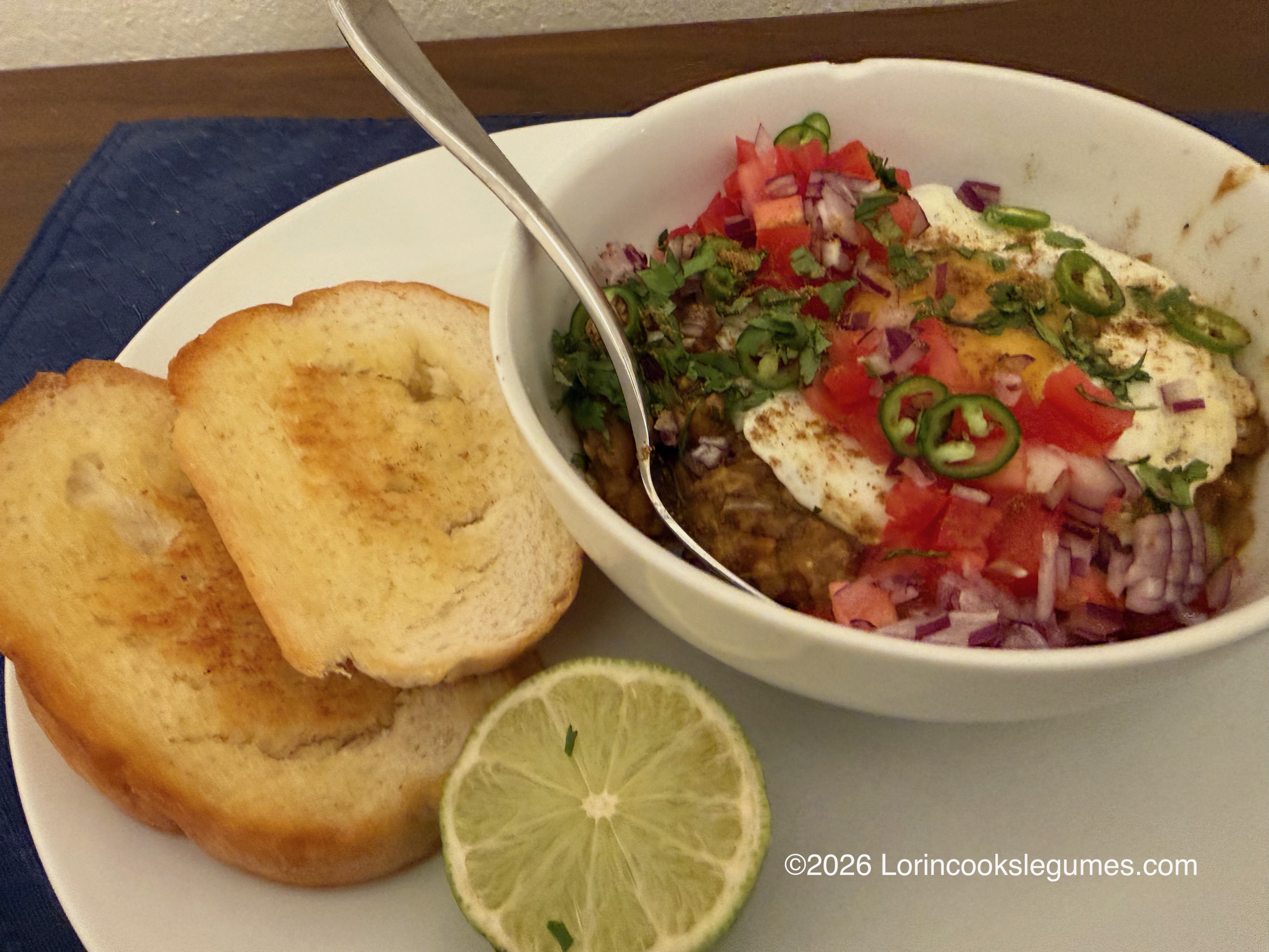 Bowl of cooked beans topped with a fried egg, chopped tomatoes, sliced green chili, onions, and cilantro served with toasted bread slices and a lime wedge on a plate
