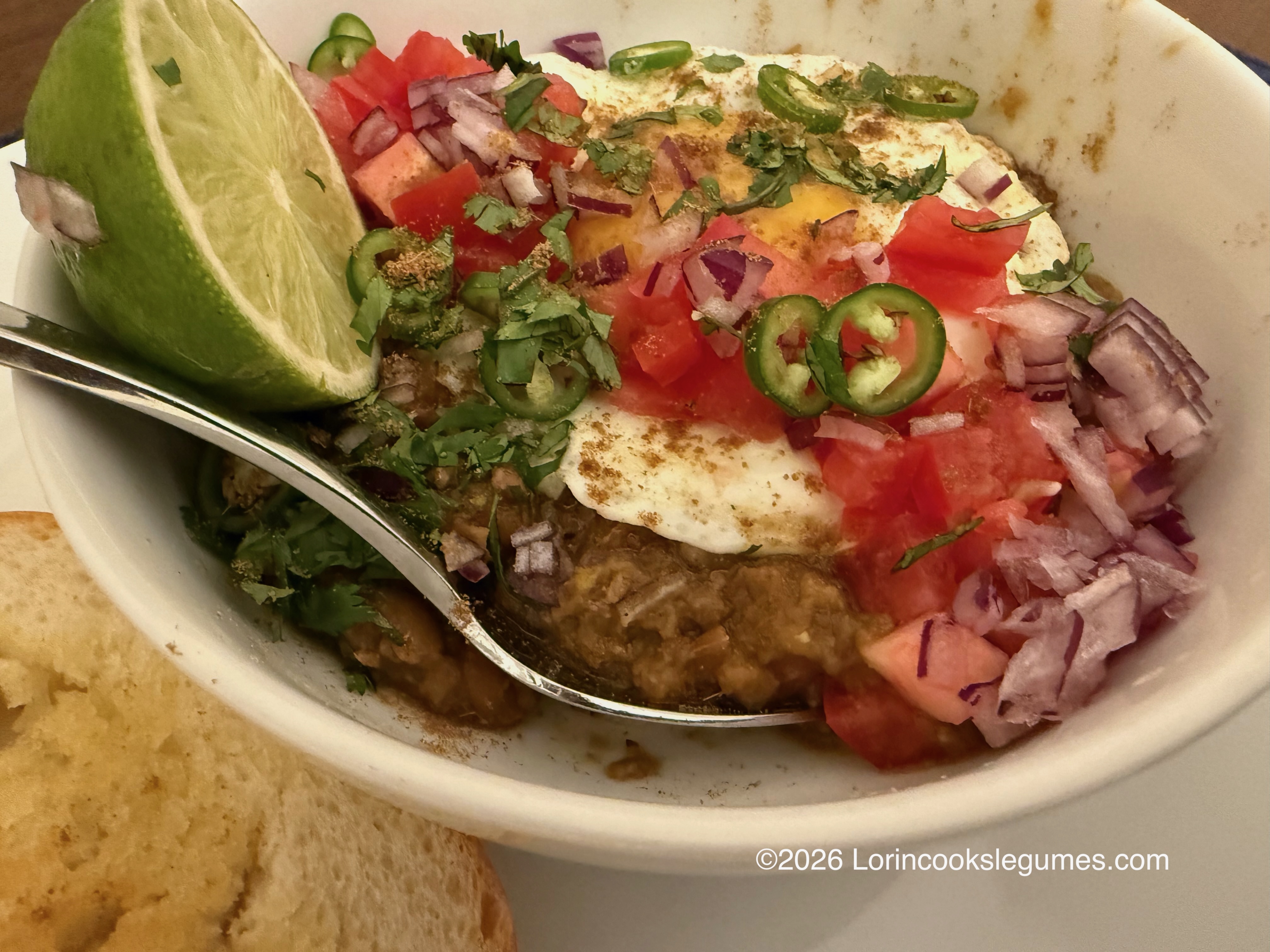 Bowl of mashed beans topped with fried egg, diced tomatoes, chopped red onions, sliced green chilies, cilantro, and a lime wedge with slices of toasted bread on the side