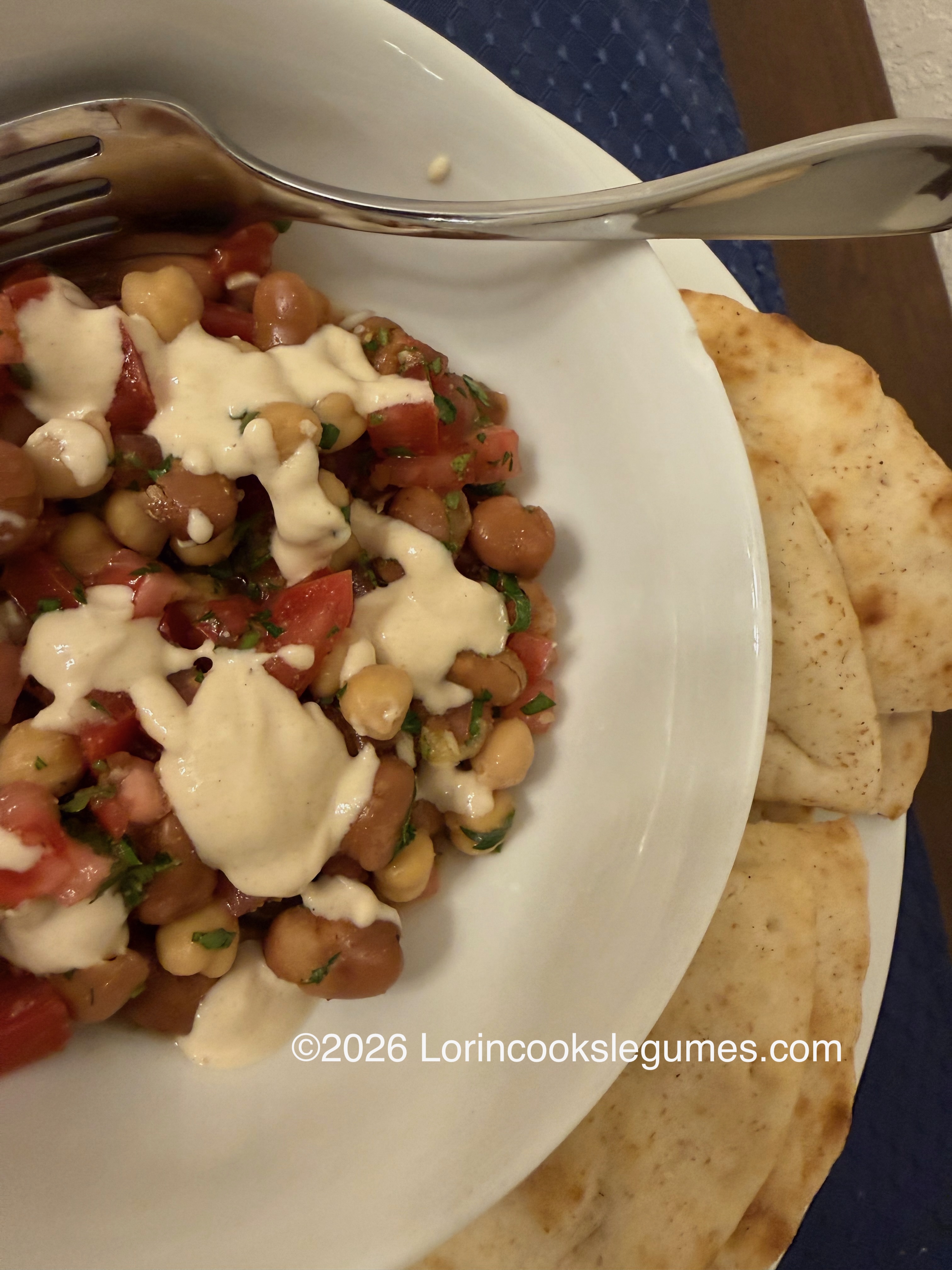 IMG_9964 Salad with fava beans, chickpeas, chopped tomatoes, and herbs drizzled with a creamy dressing, served with flatbread and a fork on a white plate