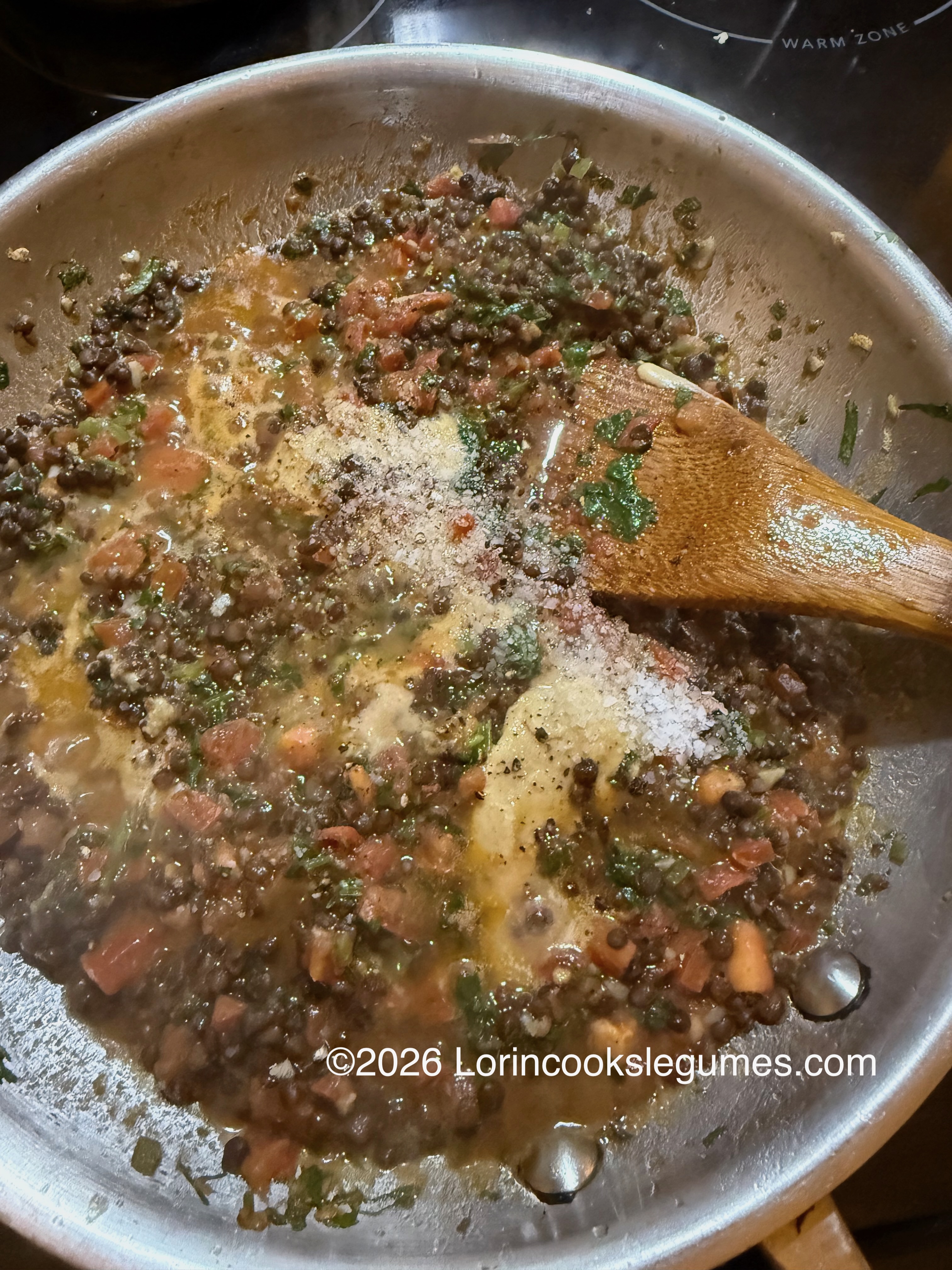 Lentils, tomatoes and herbs cooking in a pan with tahini and seasonings being stirred in with a wooden spoon