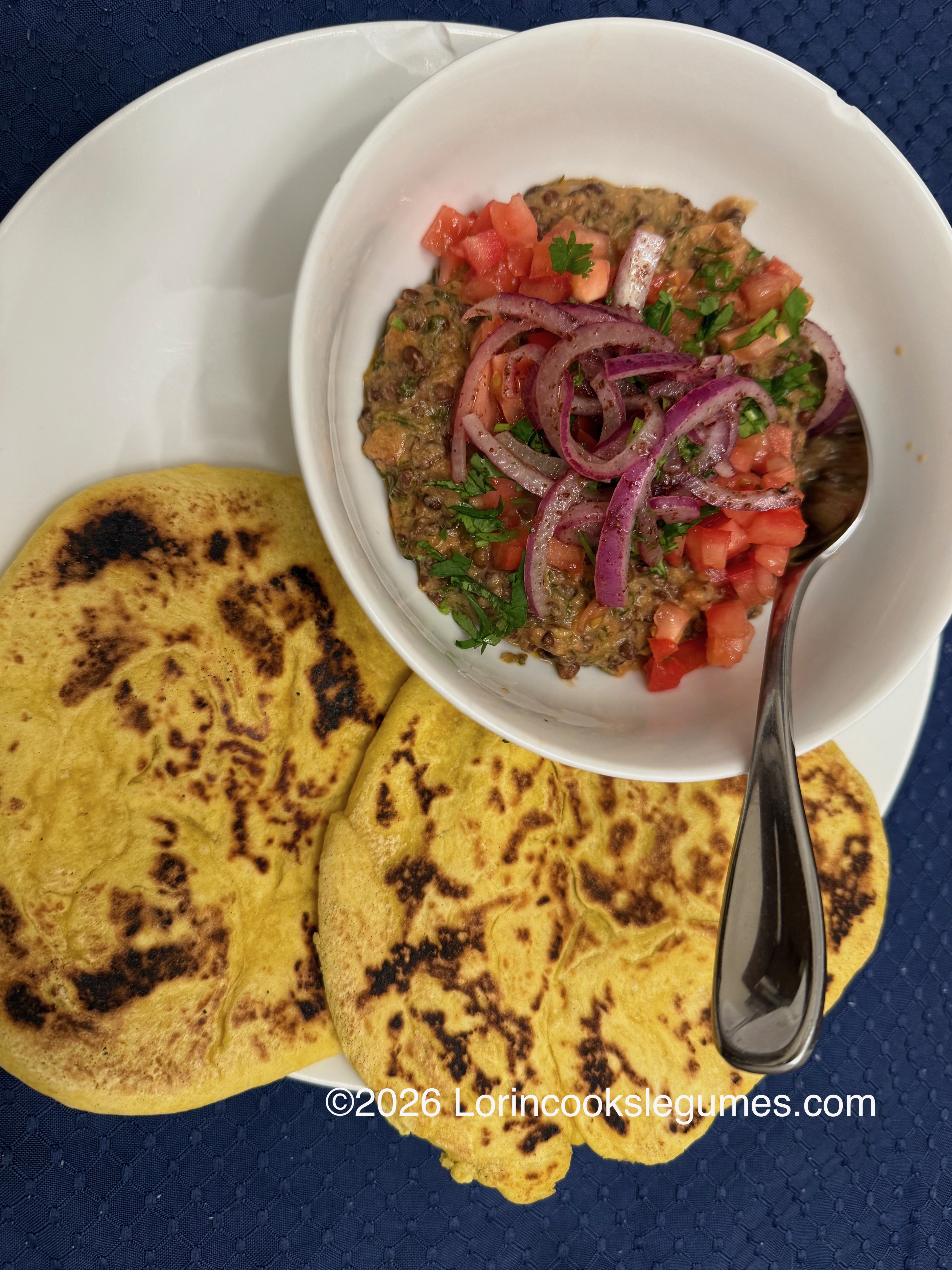 White bowl with lentil stew topped with diced tomatoes, red onions, and herbs, served with two small yellow pitas on a white plate