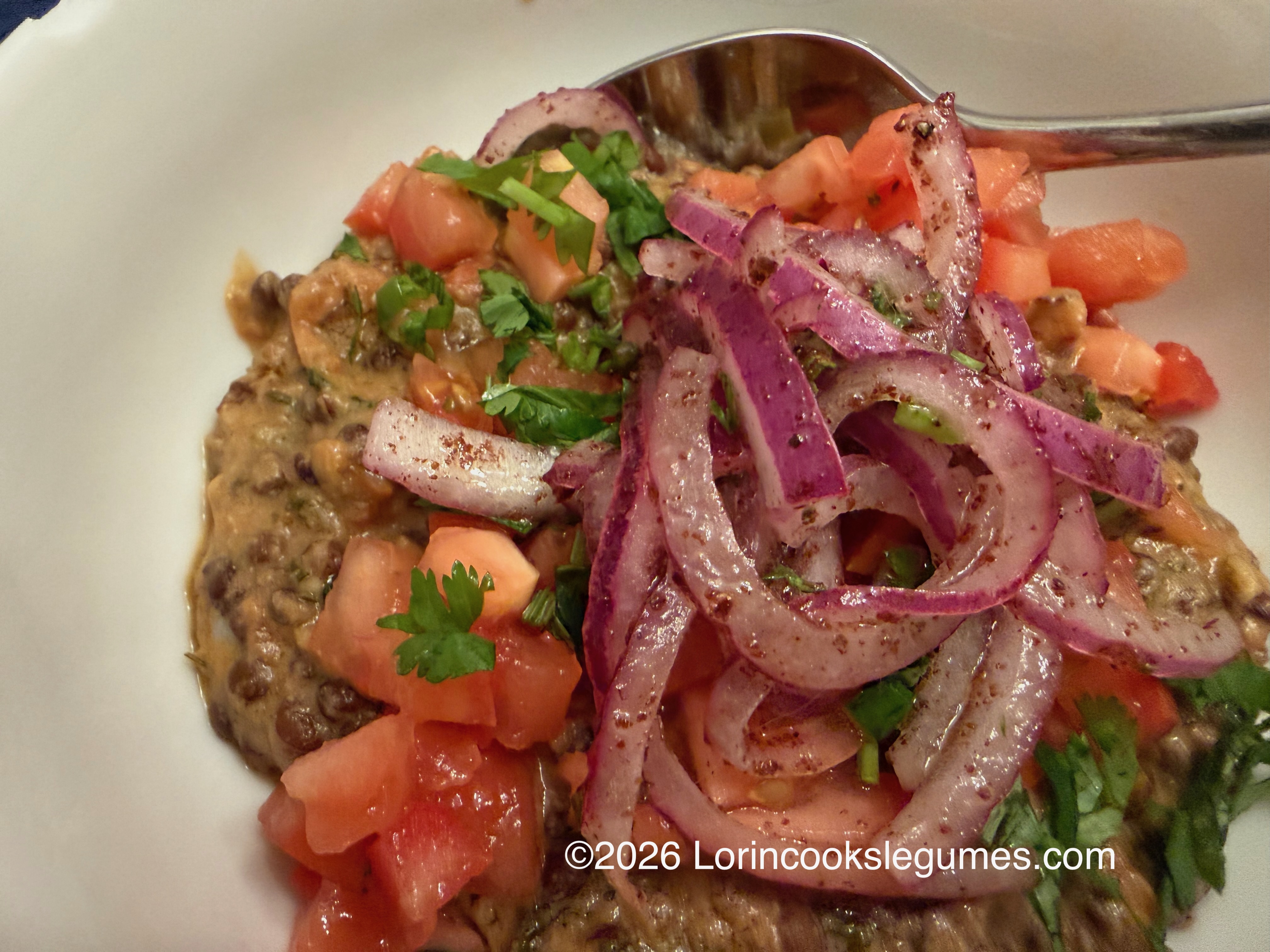 Cooked lentil dish topped with diced tomatoes, chopped cilantro, and sliced seasoned red onions with a spoon in a white bowl