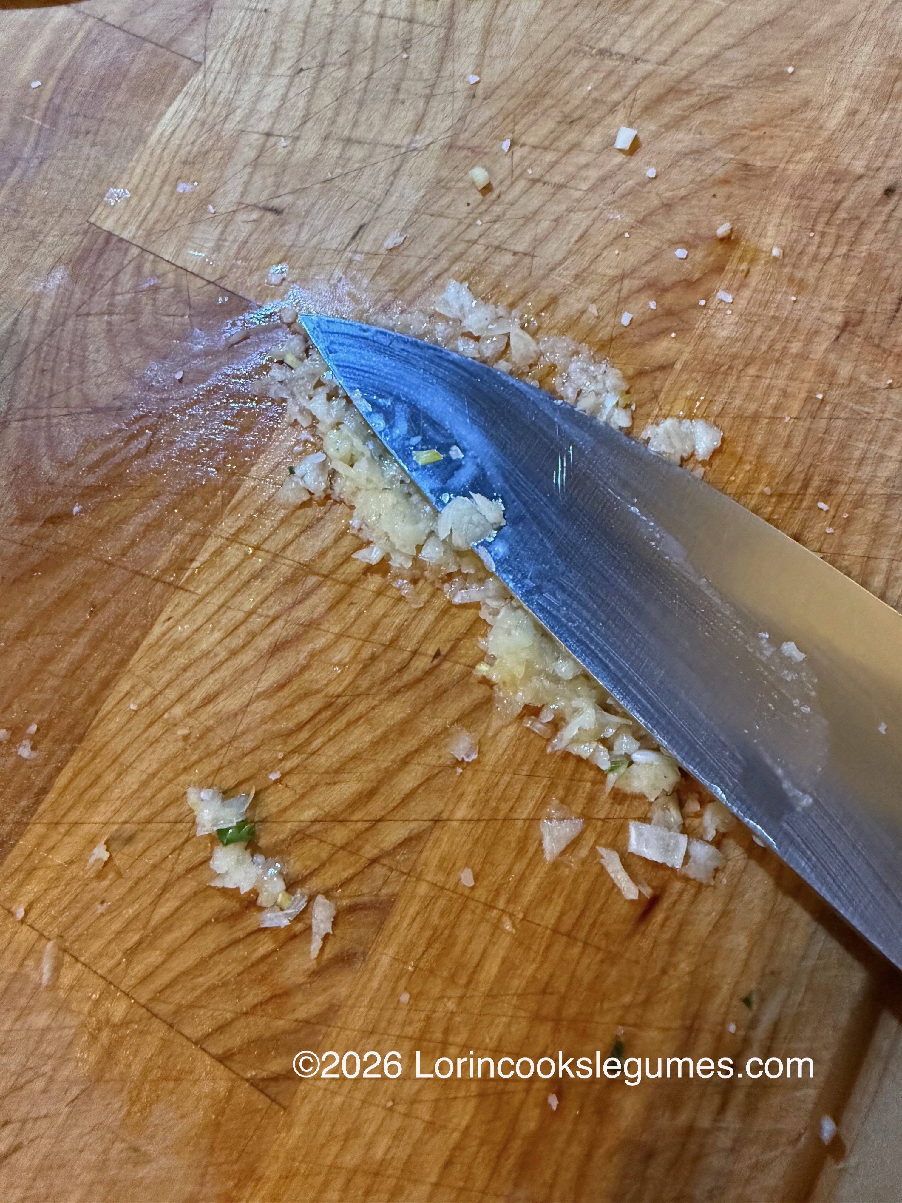 Knife crushing minced garlic on a wooden cutting board