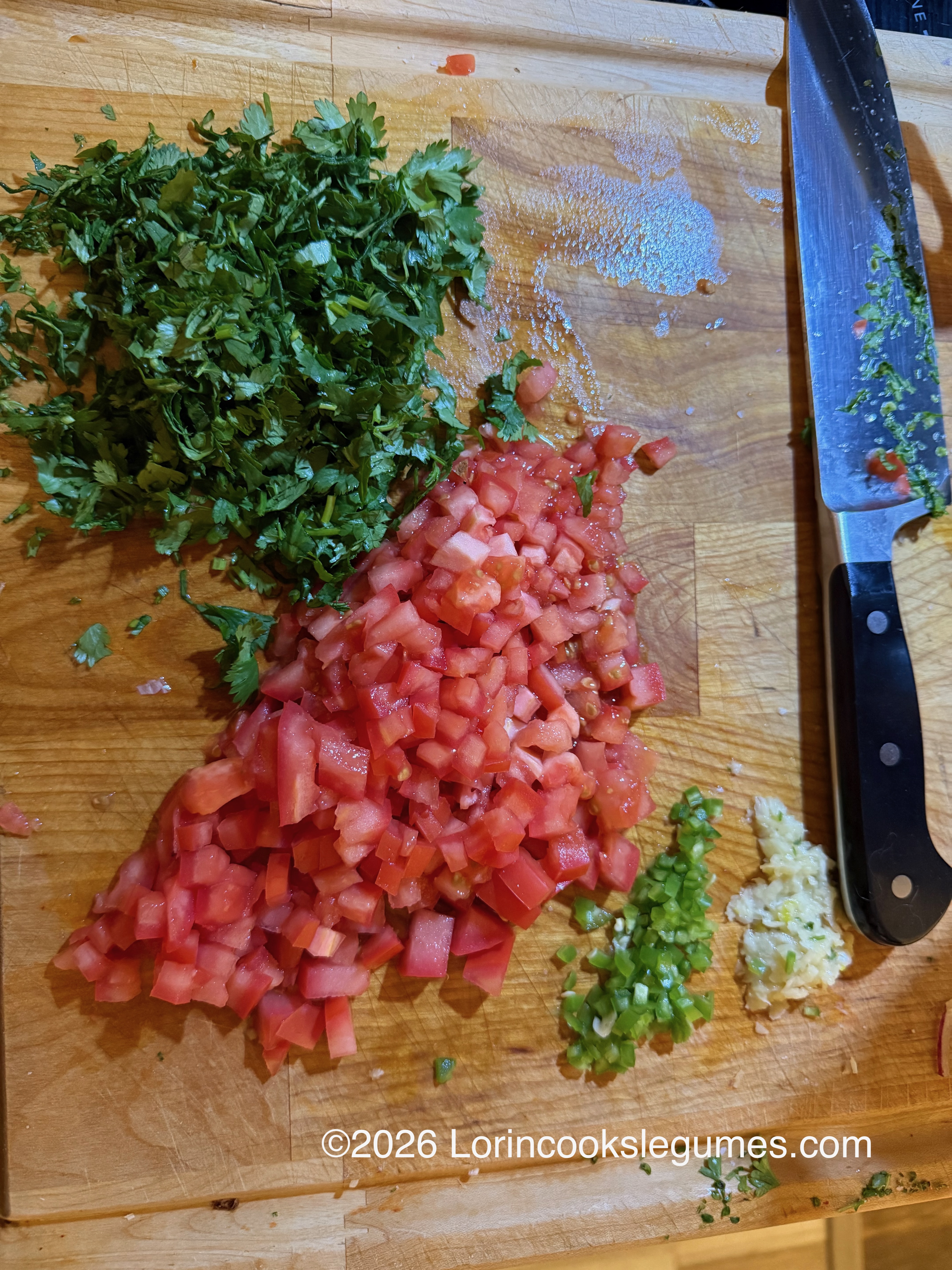 A wooden cutting board with a knife and piles of freshly chopped cilantro, tomato, green chili and garlic.