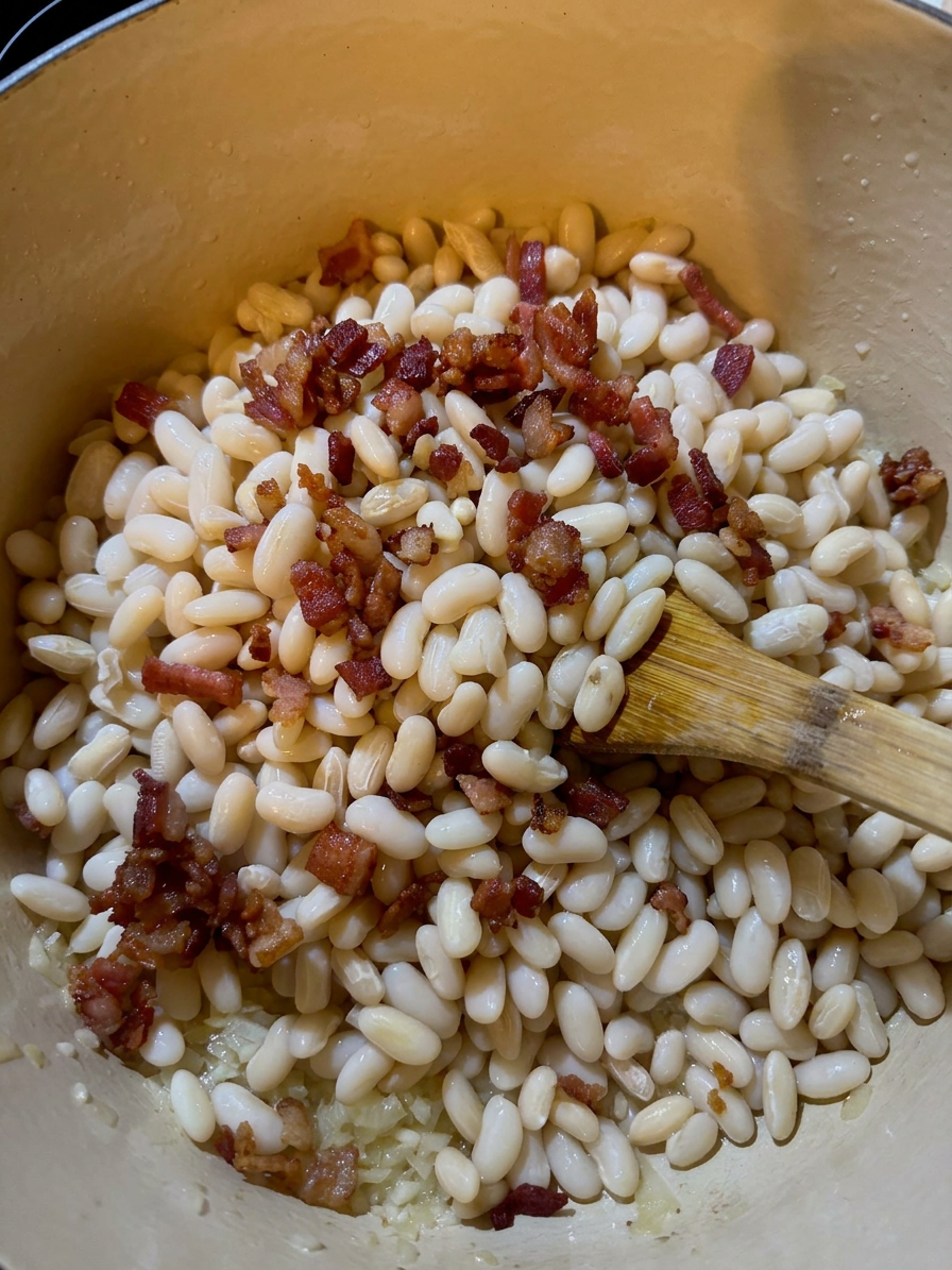 White beans being cooked in a pot with small pieces of cooked bacon and a wooden spoon