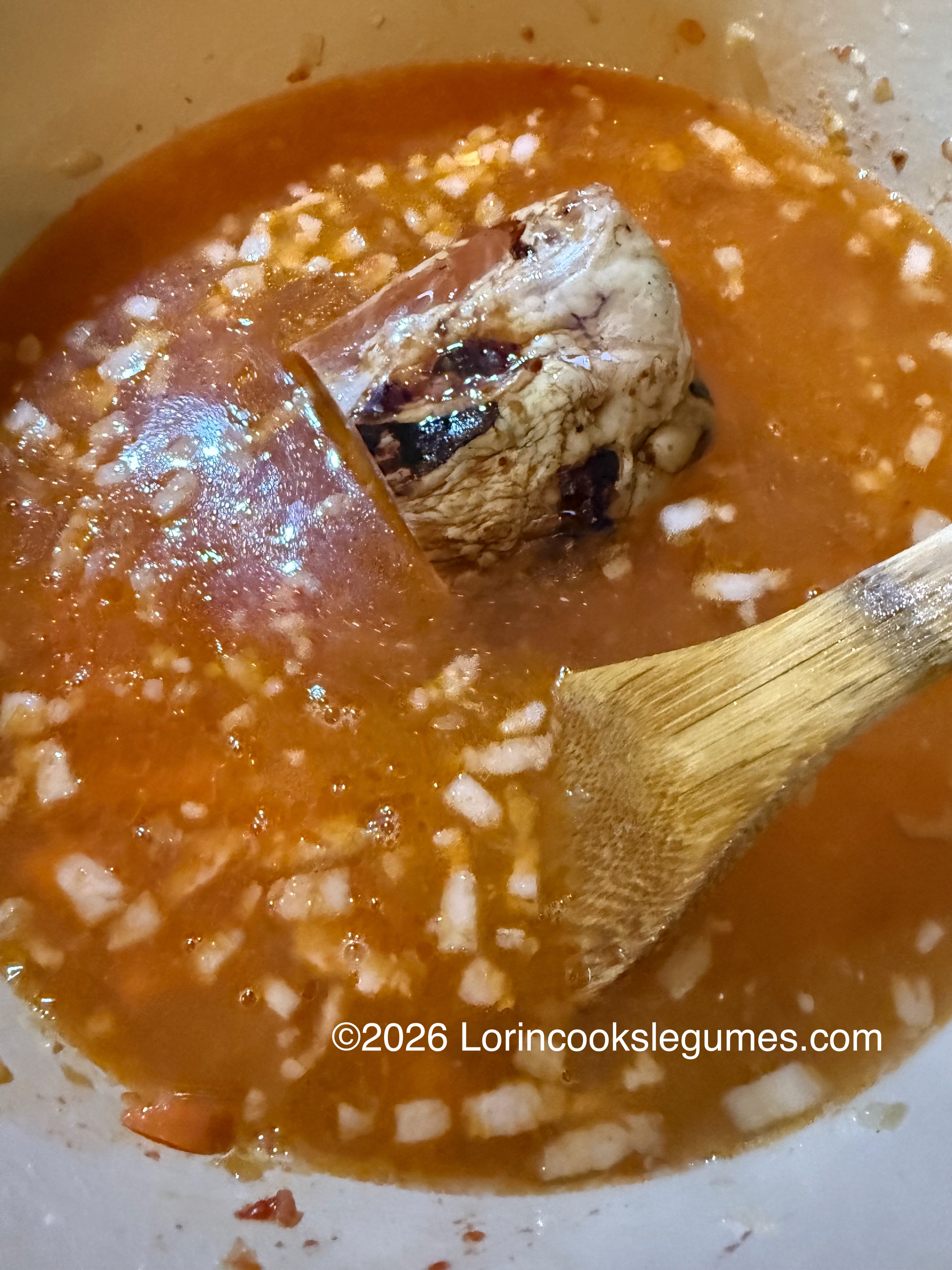 Smoked pork shank simmering in a pot of broth with chopped onions and wooden spoon