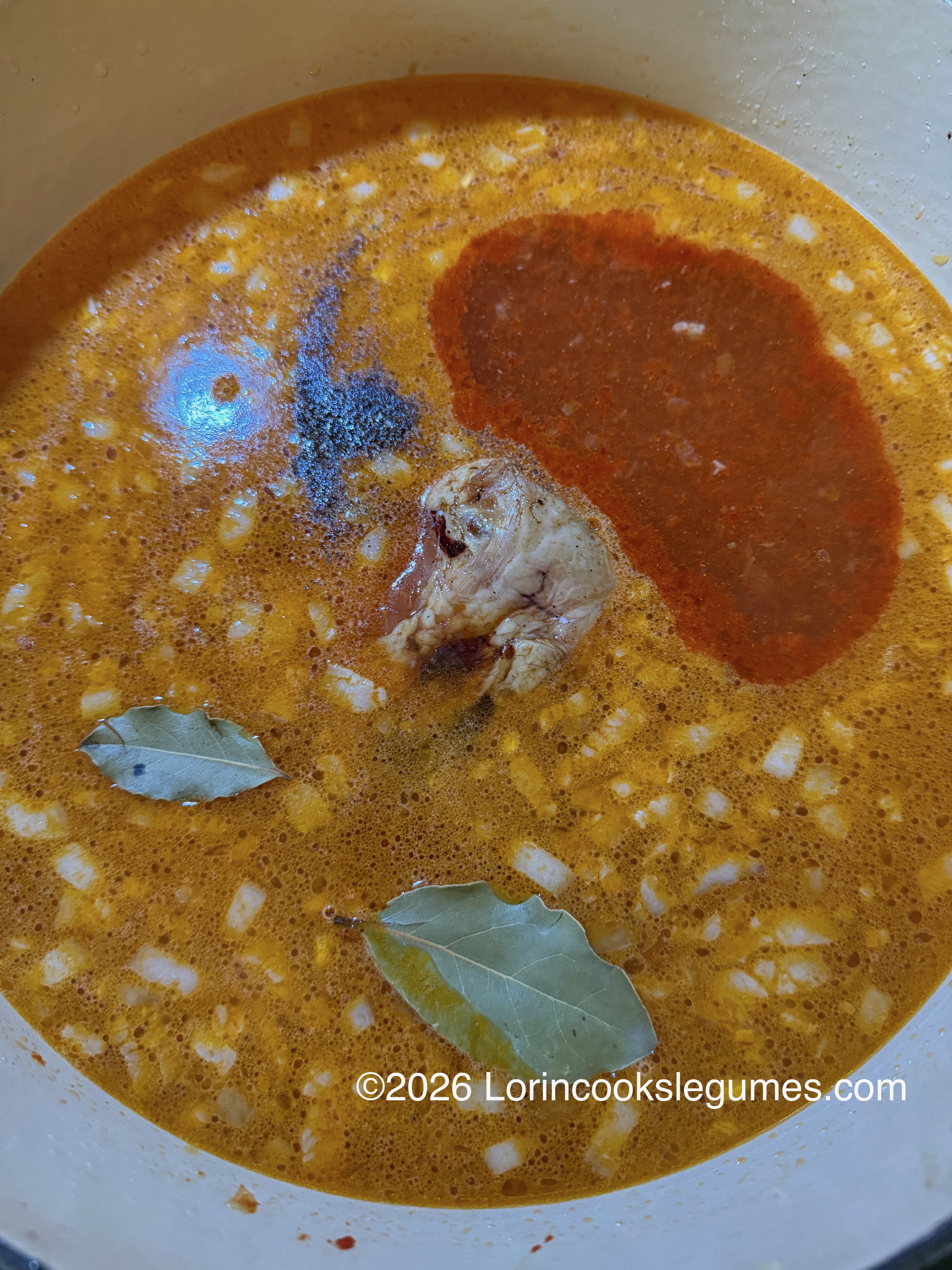 A pot of orange colored broth, containing a smoked pork shank, bay leaves, and onions, and piles of ground spices floating on the surface.
