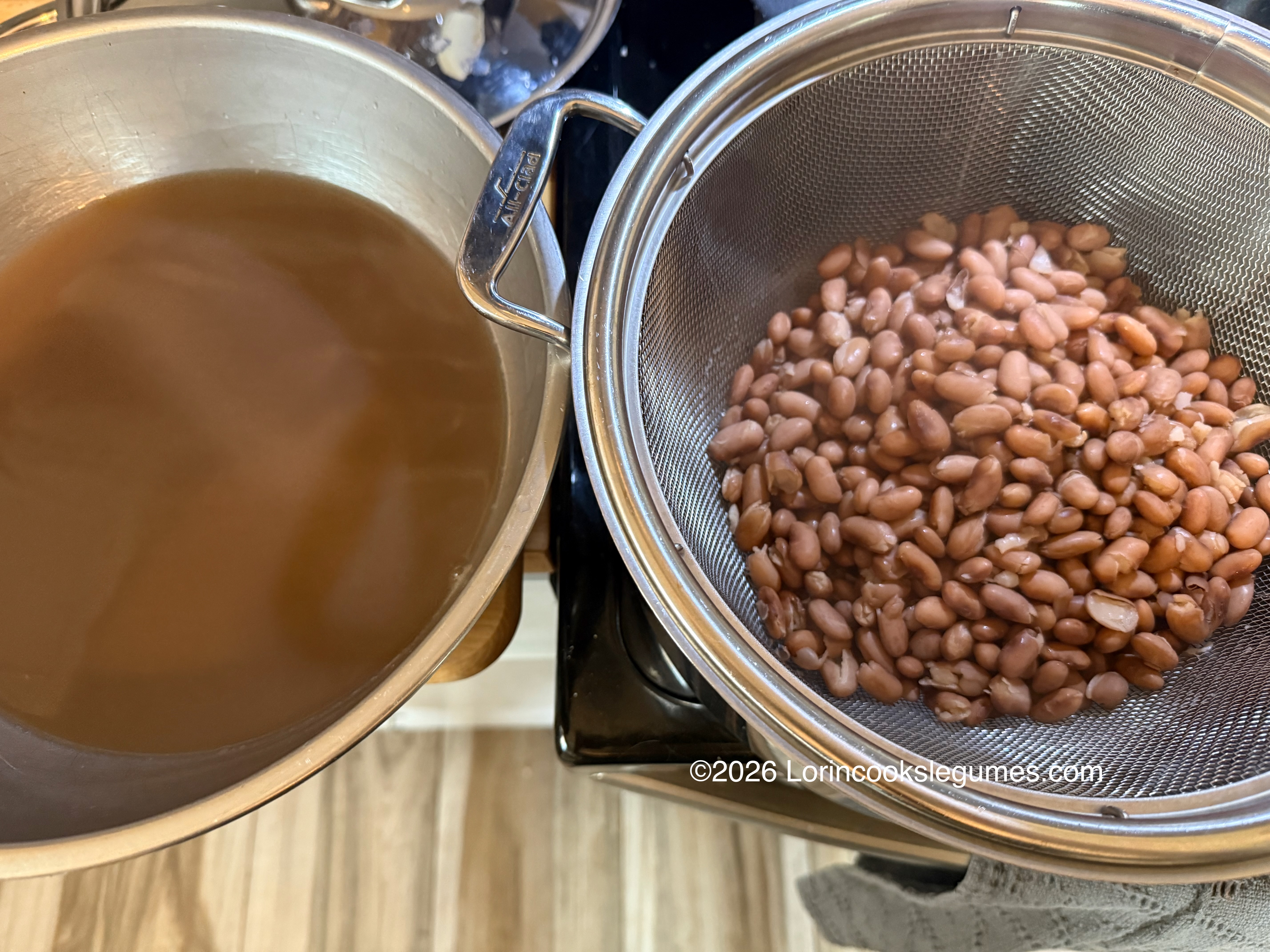 Brown beans draining in a metal colander next to a pot of brown bean broth on a stove