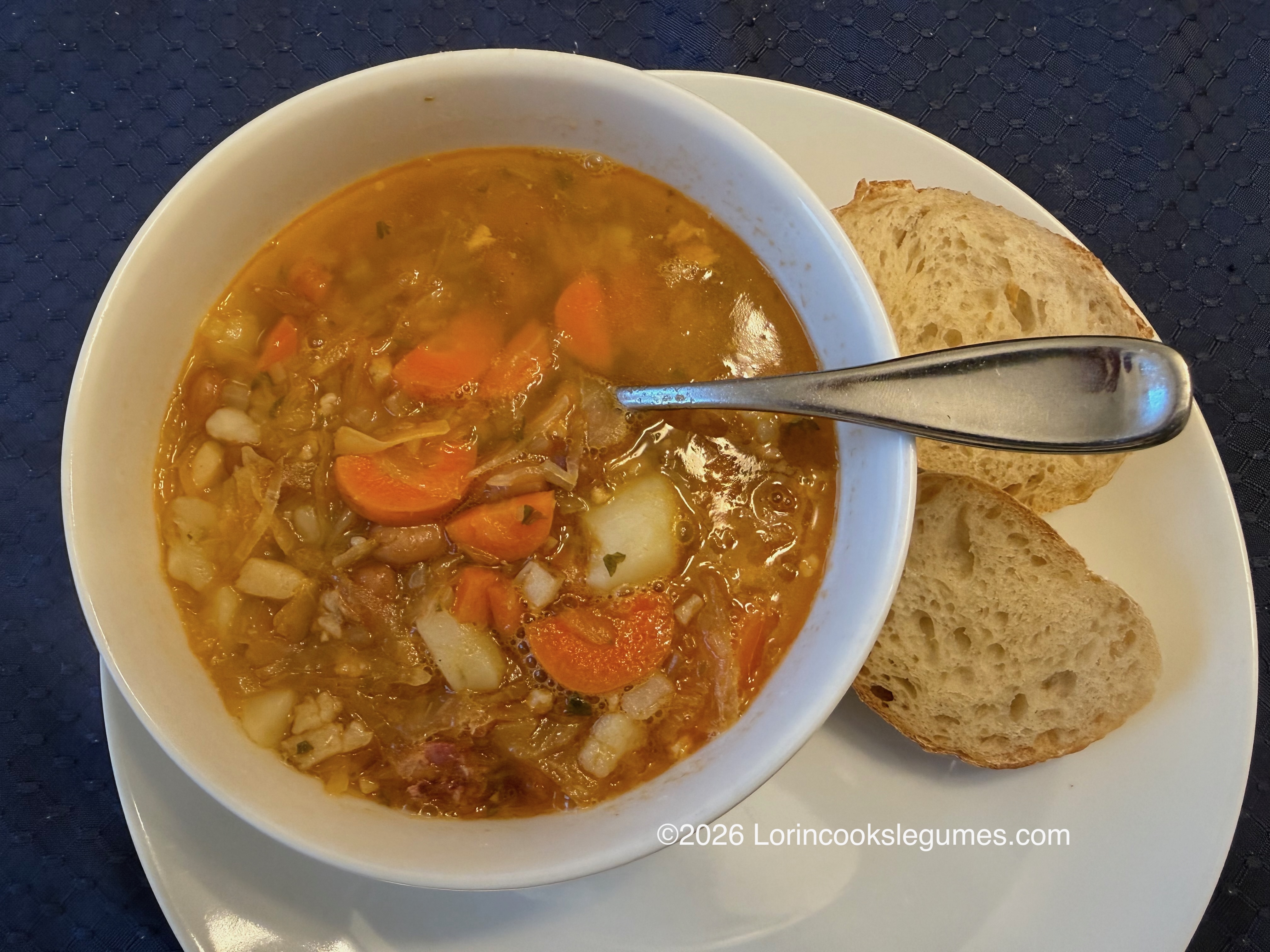 Bowl of bean soup with carrots and potatoes, served with slices of bread and a spoon on a white plate
