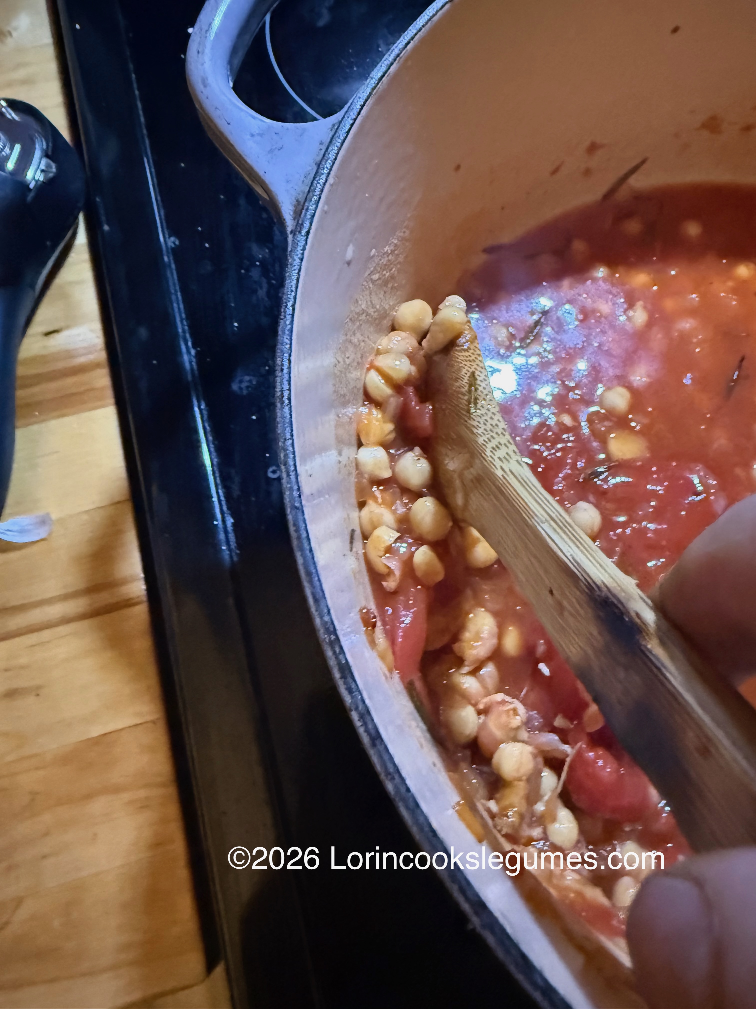 IMG_0223 A pot containing chickpeas and tomatoes with a wooden spoon mashing some of the beans against the side of the pot.