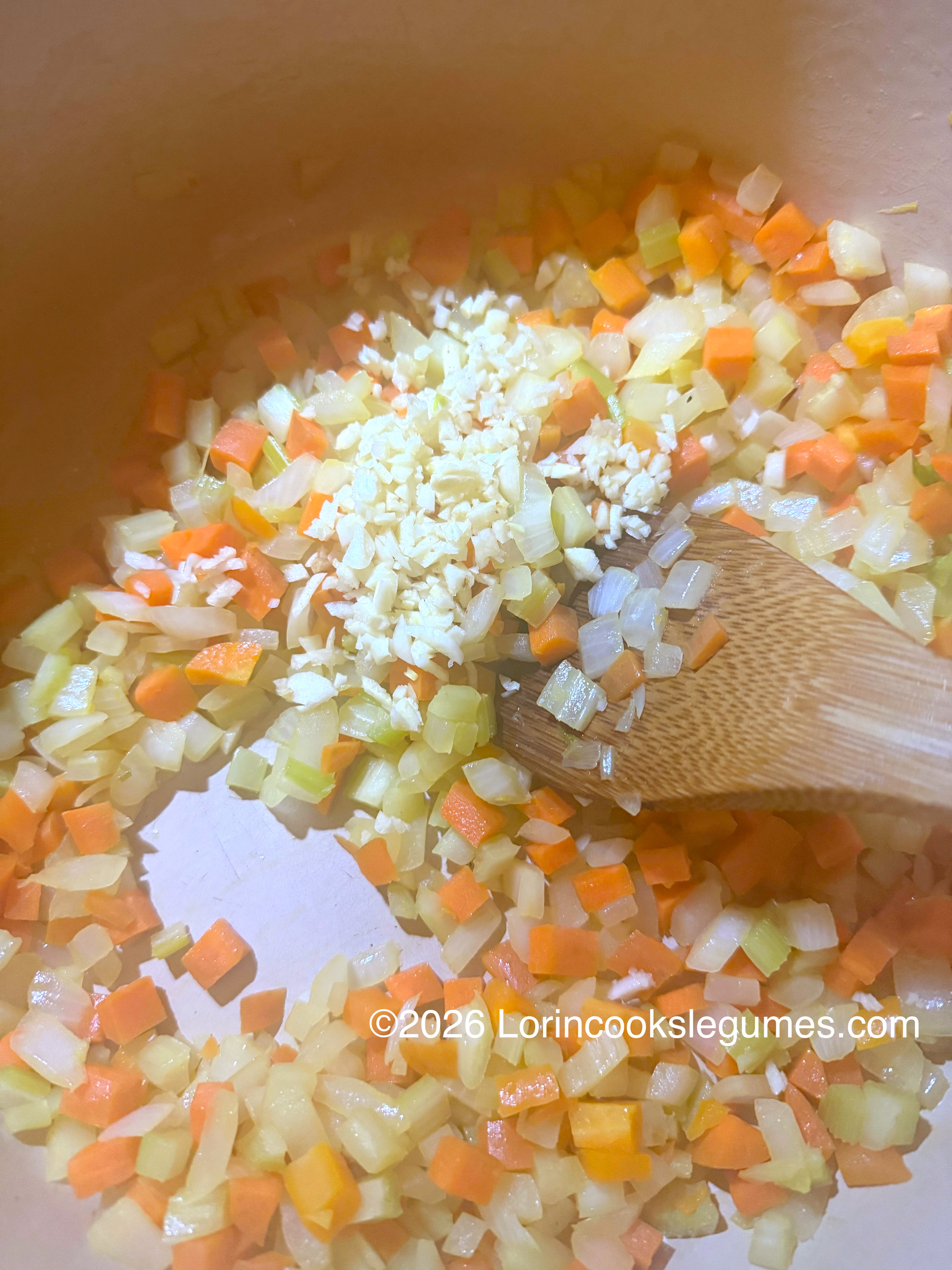 Close-up of sautéd carrots, onion and celery in a light colored pot with minced garlic and a wooden spoon.